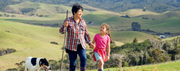 A woman with a walking stick holding hands with a young girl as both walk up a farm hill with their black and white dog.